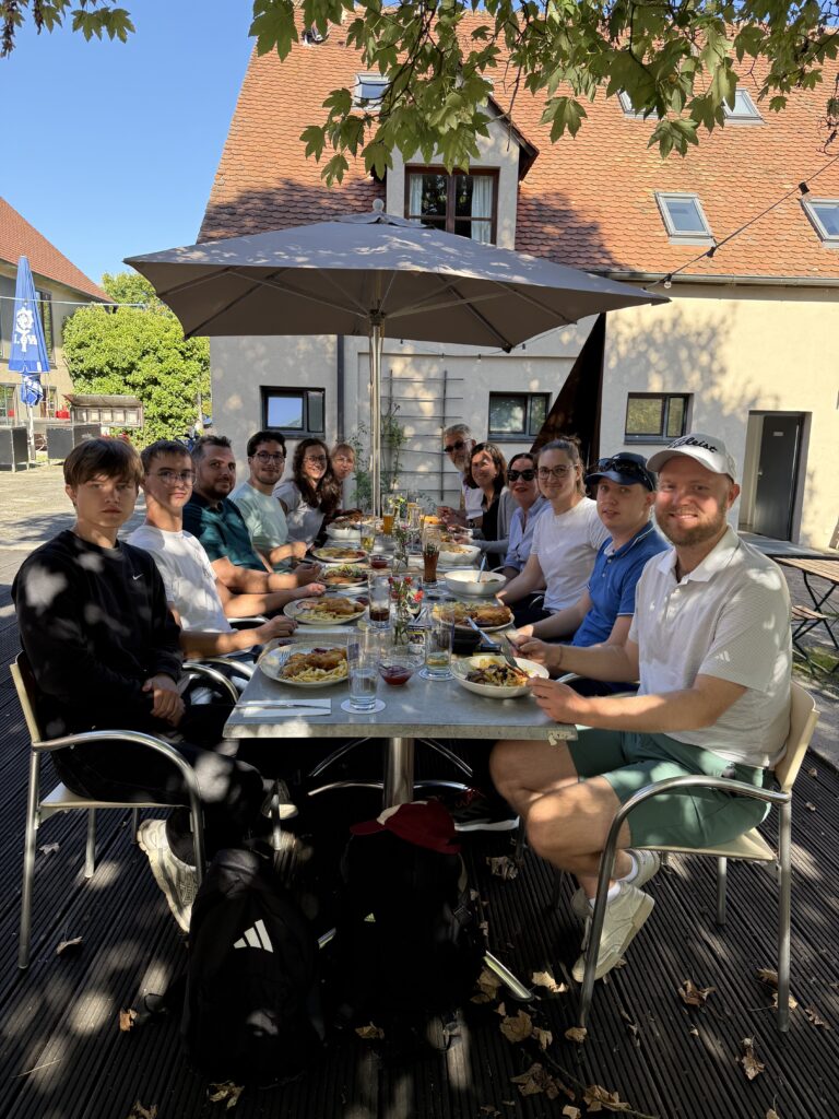Gruppe beim Essen an einem Tisch unter einem Sonnenschirm vor einem Gebäude