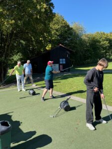 Mehrere Personen üben auf einer Golf-Übungsanlage, im Hintergrund eine Holzhütte