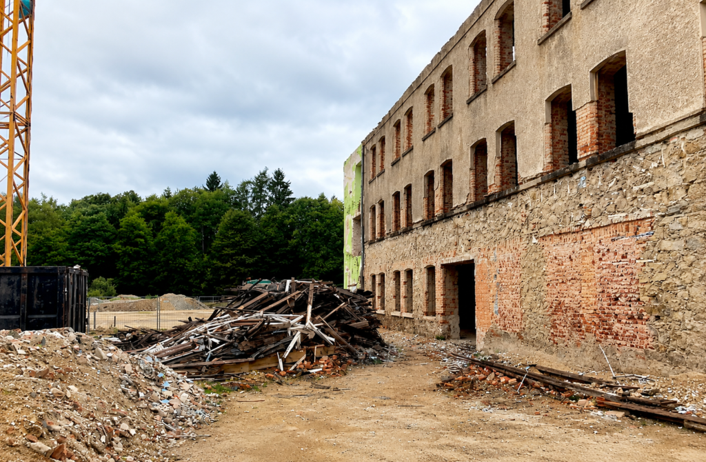 Fassade eines entkernten Bestandsgebäudes mit zugemauerten Öffnungen, davor Abbruchmaterial, links ein Baukran