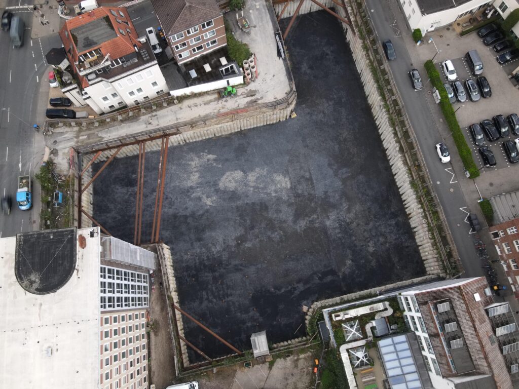 Senkrechter Blick von oben auf die rechteckige Baugrube zwischen Straße, Parkplatz und Nachbargebäuden