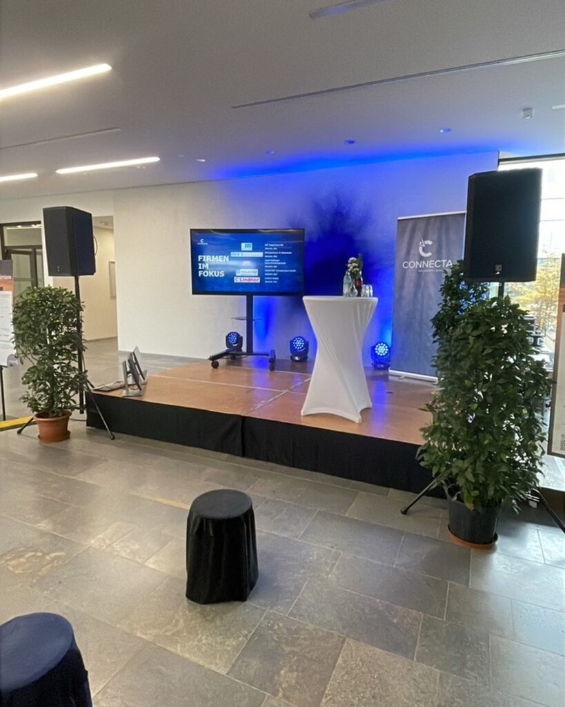 Leere Vortragsbühne der CONNECTA im Foyer mit Bildschirm, Stehtisch und blauer Beleuchtung