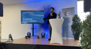 Ein MKI-Mitarbeiter hält auf der Bühne der CONNECTA einen Vortrag, auf dem Bildschirm daneben steht „Komplexes kreativ meistern.“